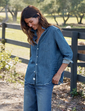 A person with long brown hair stands outdoors, facing away, next to a black wooden fence with trees and grass in the background, wearing the FRANK & EILEEN EILEEN Relaxed Button Up Shirt in Famous Denim Distressed Indigo Wash.