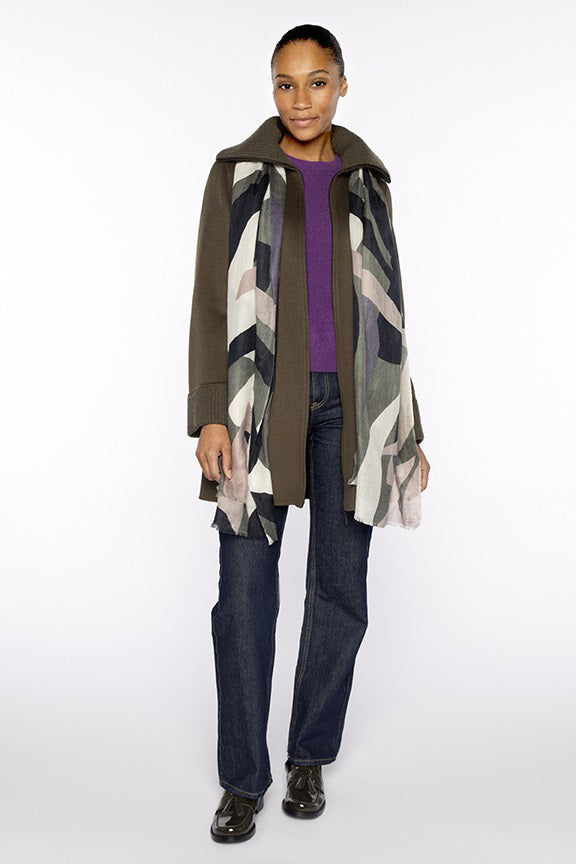 A woman faces forward, smiling slightly with her hair pulled back. She wears a dark olive coat, purple top, blue jeans, black shoes, and the KINROSS CASHMERE ABSTRACT PRINT SCARF in neutral tones against a white background.