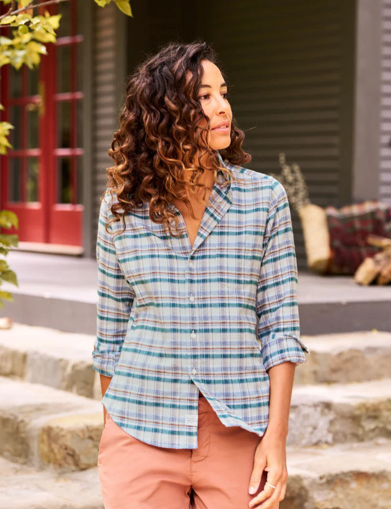 A woman with curly hair smiles and looks up as she stands outside next to a tree, wearing the Frank &amp; Eileen Barry Tailored Button Up in Cream Green Blue Plaid Italian Flannel and rust-colored pants. Her hands are in her pockets, and a house with a red door and a pile of firewood is visible in the background.