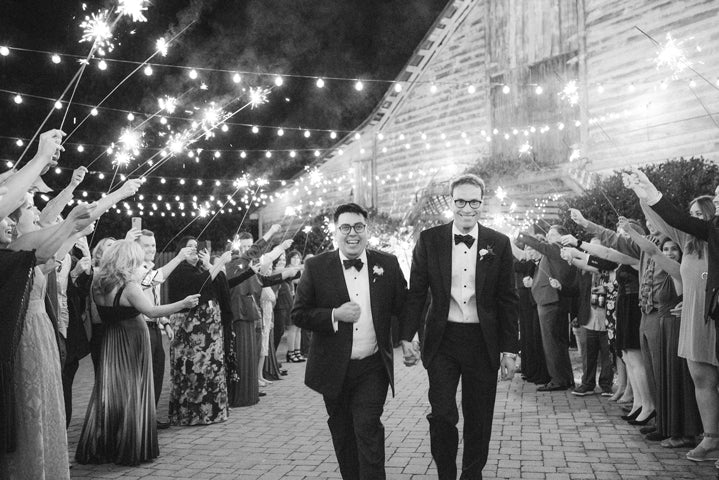 Two grooms in tuxedos walk hand-in-hand, smiling, under string lights and sparklers held by cheering guests, outside a rustic barn at night. The scene is joyful and celebratory.