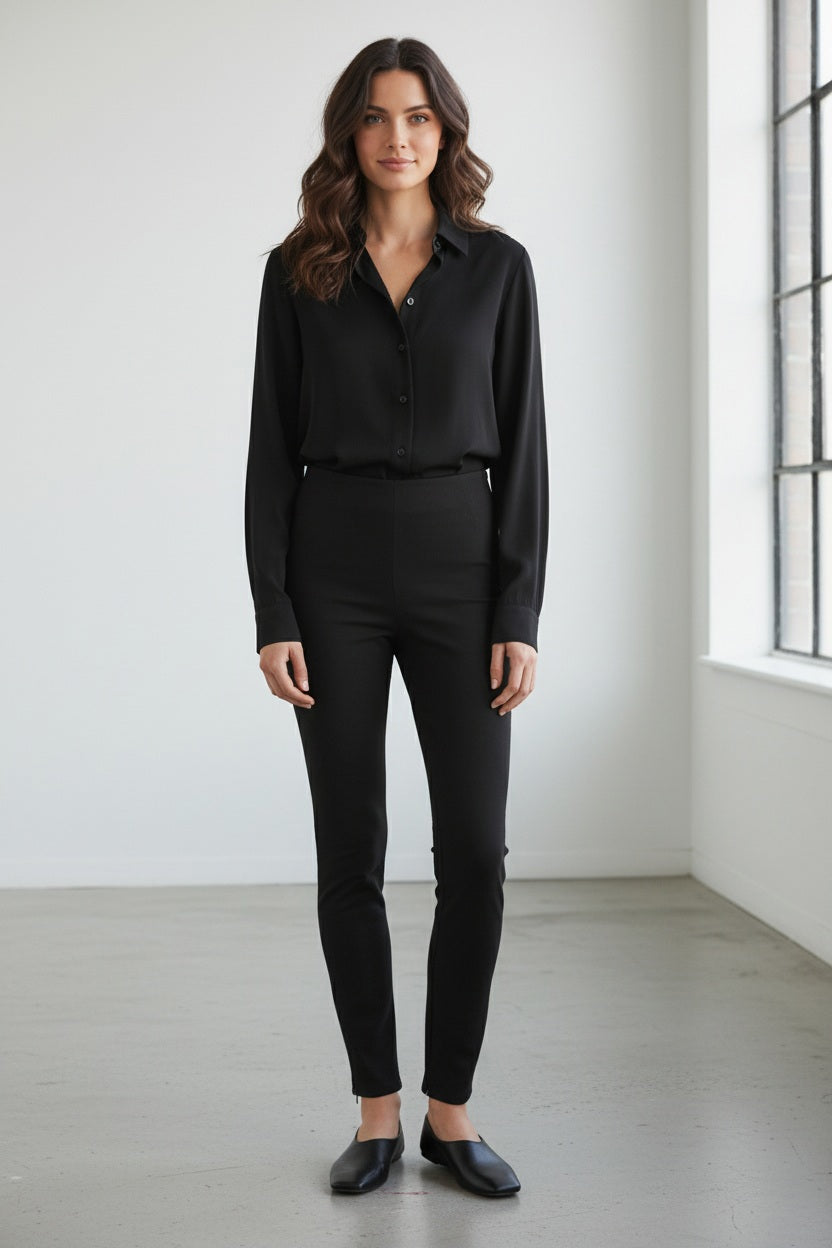 A woman with wavy brown hair stands indoors near a large window, wearing the MEIMEIJ Legging Pant with Zipper at Ankle in Black by MEIMEIJ. She pairs the sleek black pants with a button-up shirt and black shoes, looking at the camera with a neutral expression.