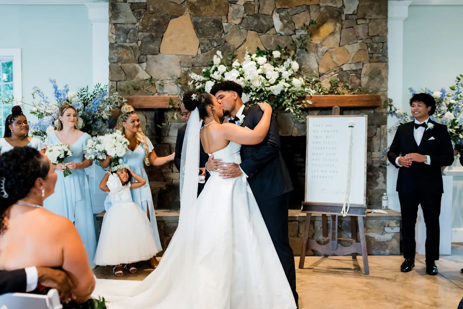 A bride and groom share a kiss at their wedding ceremony, surrounded by bridesmaids in light blue dresses and groomsmen in tuxedos, with a flower girl smiling nearby. White flowers and a stone fireplace decorate the background.