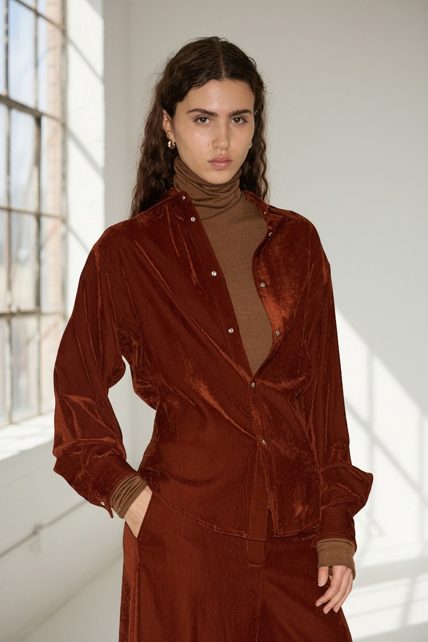 With long wavy brown hair, a person exudes Italian elegance wearing NUDE’s fitted shirt in tobacco layered over a brown turtleneck and paired with matching pants, as they stand indoors beside a sunlit window.