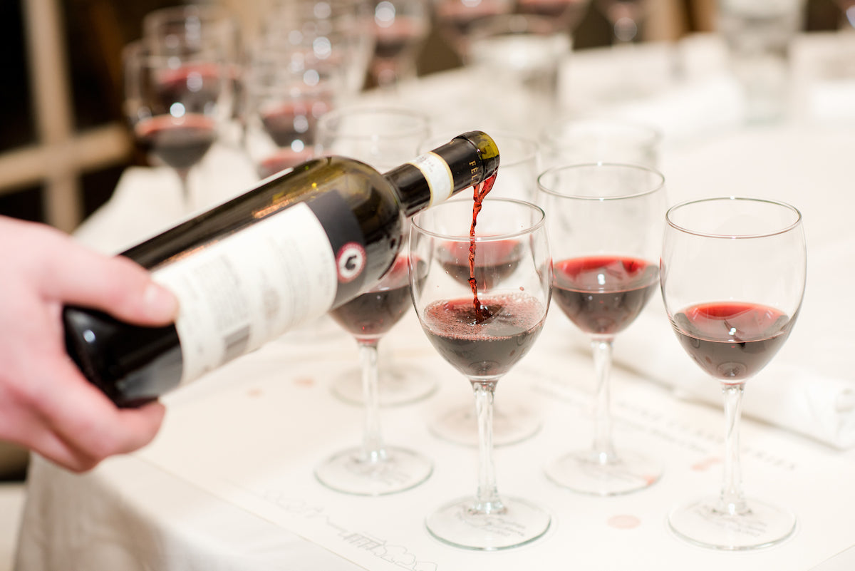 A hand pours red wine from a bottle into a glass, surrounded by several other partially filled wine glasses on a white tablecloth.