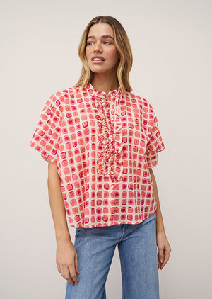 A woman wears the DAISY BLOUSE IN PINK PRINT by 0039 ITALY, featuring pink and red square patterns with ruffles, paired with light blue jeans against a plain light background.
