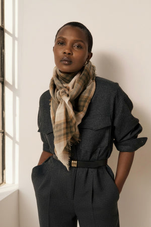 A woman with short hair stands against a plain light background, looking directly at the camera with a neutral expression. She wears a dark belted dress and the HARTFORD Checked Wool Scarf in Taupe, made in India.
