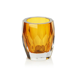 A faceted amber-colored glass vase—the LA BOHEME HANDMADE POLISHED CUT GLASS VASE / HURRICANE in Amber by ZODAX—rests on a wooden surface, accompanied by a blurred book, a matching glass piece, and a small gold pump dispenser.