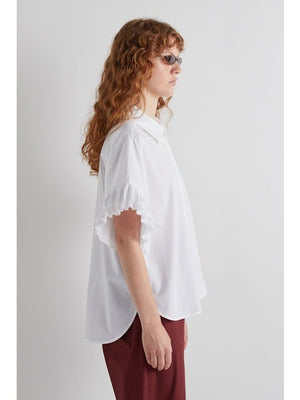 A person with long, wavy hair and sunglasses models the MEIMEIJ - WHITE COTTON SHIRT, featuring ruffled sleeves, paired with maroon pants, standing in profile against a plain white background.