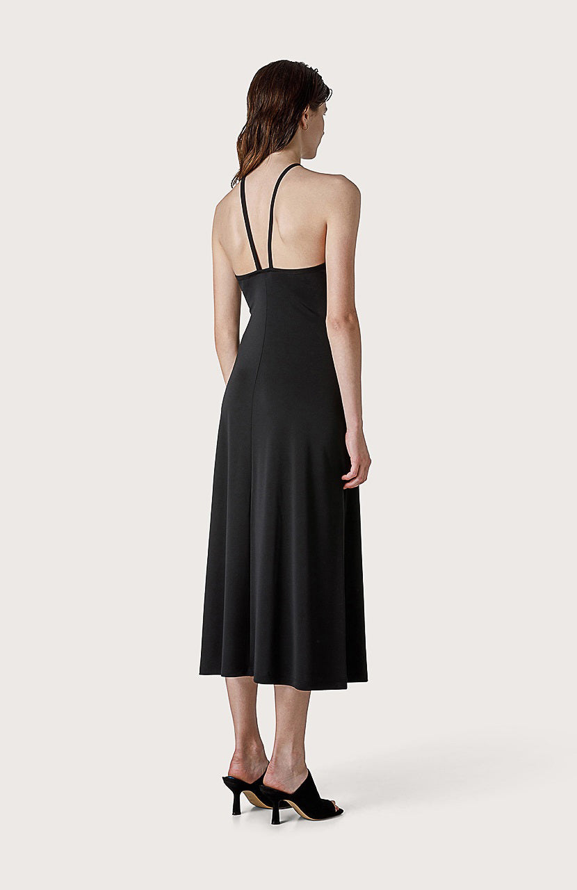 A woman stands against a plain background wearing the SEVENTY JERSEY DRESS, which is a black, sleeveless, ankle-length party dress with thin straps crossing at the neck. Her hair is wet and she is wearing black open-toed shoes. She has a neutral expression and her arms are relaxed by her sides.