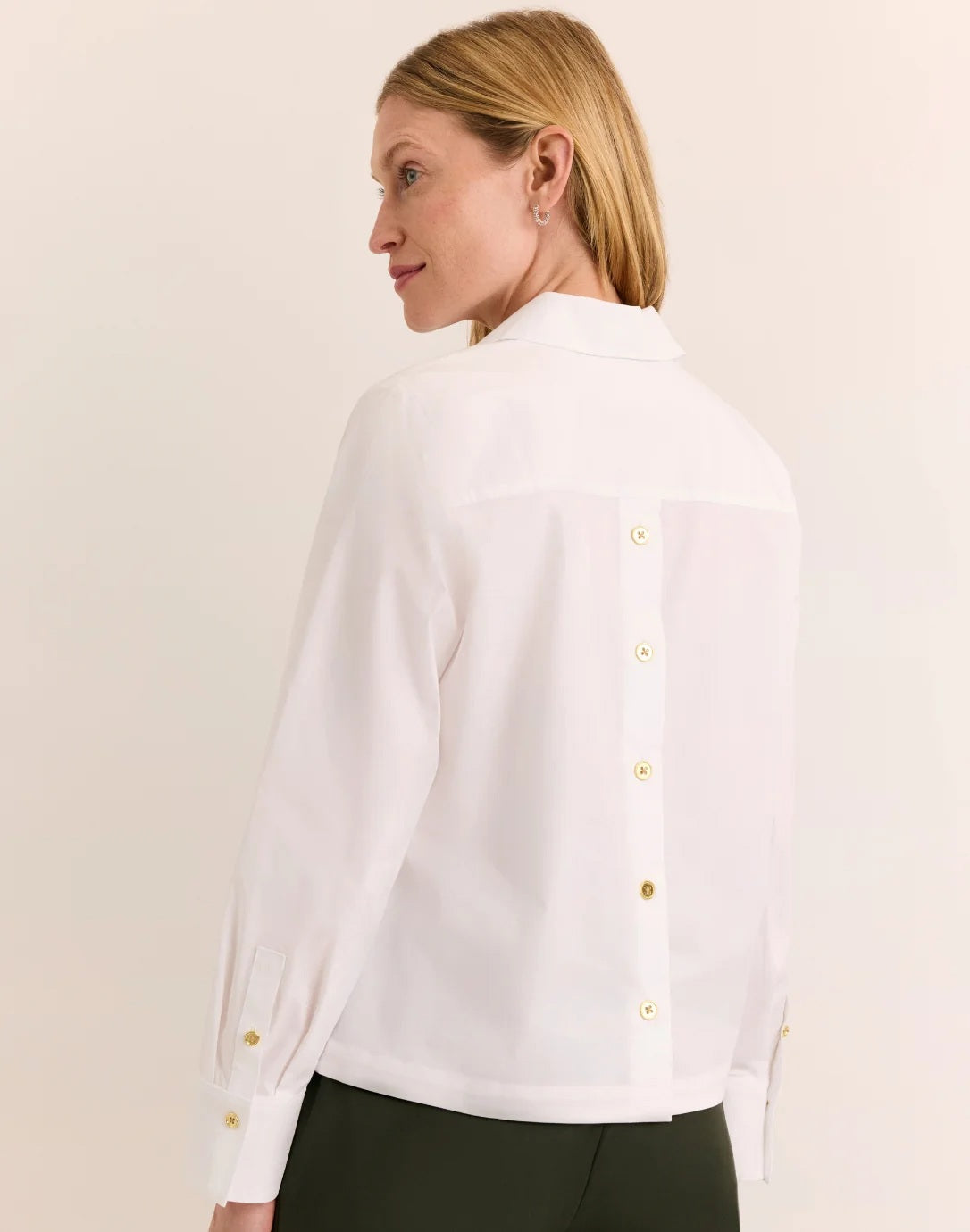 A woman with long blonde hair models the HINSON WU Monica Long Sleeve Button Back Top in white—a collared stretch polished cotton blouse featuring a split neckline and long sleeves—paired with dark green pants, standing against a plain light background.