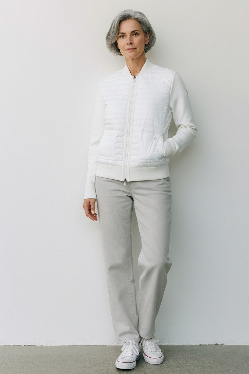 A woman with short gray hair stands against a white wall wearing the SAVE THE DUCK BRIONY LIGHTWEIGHT PUFFER—a cropped, quilted white jacket—paired with light gray pants and white sneakers. An ideal layering option for cooler days.
