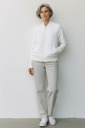 A woman with short gray hair stands against a white wall wearing the SAVE THE DUCK BRIONY LIGHTWEIGHT PUFFER—a cropped, quilted white jacket—paired with light gray pants and white sneakers. An ideal layering option for cooler days.