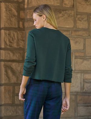 A woman with wavy dark hair stands by large windows, smiling as she wears the FRANK & EILEEN Jackie Sweatshirt Cardigan in Triple Fleece Pine from FRANK & EILEEN, paired with blue plaid pants and black loafers. One hand is tucked into her pocket and her legs are crossed.