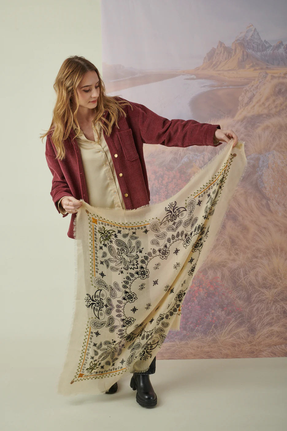 A young woman with light brown hair wears the STORIATIPIC Nelly Square Cashmere and Wool Embroidered Bandana Scarf, paired with a light gray top, standing before a softly colored nature-themed backdrop.
