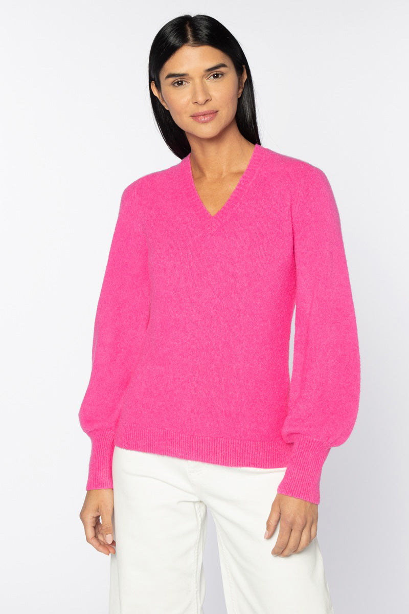 A woman with long black hair is standing against a white background. She is wearing a bright pink KINROSS Cashmere Stretch Boucle Vee Neck Sweater, which features balloon sleeves, and a ribbed hem and cuffs. Accompanying her stylish top are white pants. The sweater&#39;s soft cashmere material adds to her relaxed look, with her arms resting naturally by her sides and a neutral expression on her face.