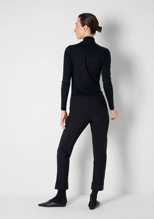 A person stands facing away from the camera against a plain light background, styled in a fitted black turtleneck and KATHARINA HOVMAN - SIMPLE PANTS by KATHARINA HOVMAN, which feature stretch quality for added comfort. Their hair is tied back in a neat, low bun, and black shoes complete the minimalist look.