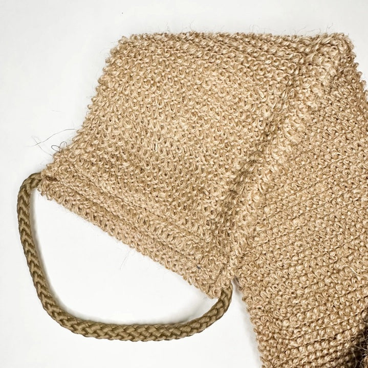 A close-up of the SMILE BOUTIQUES NATURAL ORGANIC JUTE BACK SCRUBBER, featuring a brown, woven jute design with a braided handle at one end and laid flat on a white background—this handmade bath accessory offers a sustainable upgrade to your shower routine.