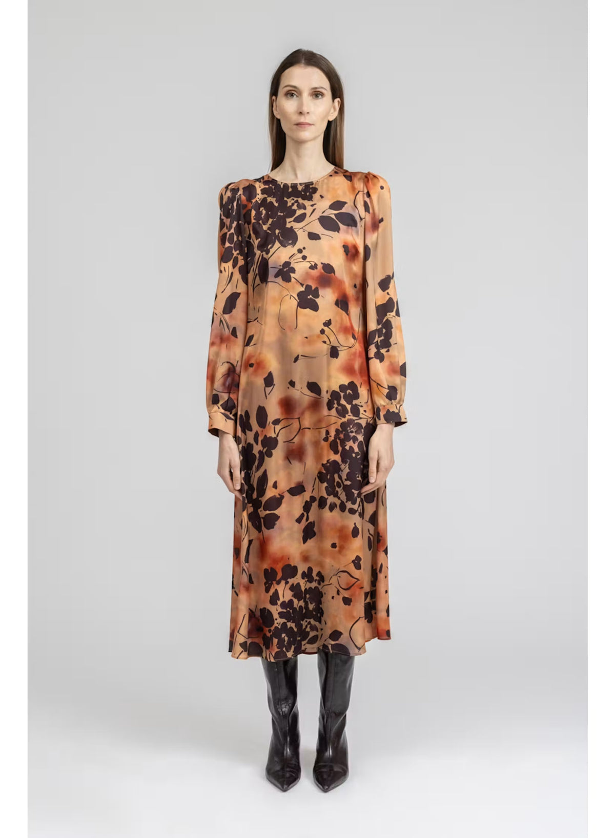 A woman stands with her back to the camera, wearing the MILVA MI - BURNT ORANGE PRINTED DRESS by MILVA—a long-sleeved, viscose midi dress in a rich brown shade accented with an orange and black botanical print—paired with black knee-high boots against a plain light background.