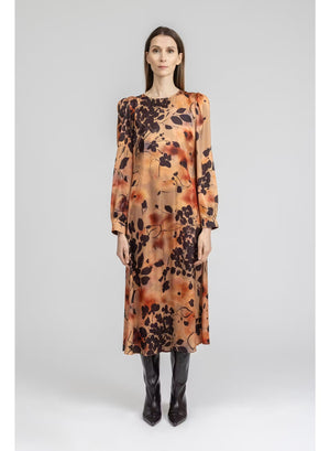 A woman stands with her back to the camera, wearing the MILVA MI - BURNT ORANGE PRINTED DRESS by MILVA—a long-sleeved, viscose midi dress in a rich brown shade accented with an orange and black botanical print—paired with black knee-high boots against a plain light background.