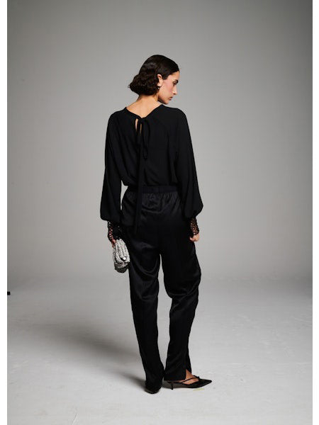 A woman with short dark hair faces away, wearing the MILVA MI - CROCHET CUFF BLOUSE by MILVA, black pants, and pointed black shoes. She holds a silver textured clutch against a plain gray background.