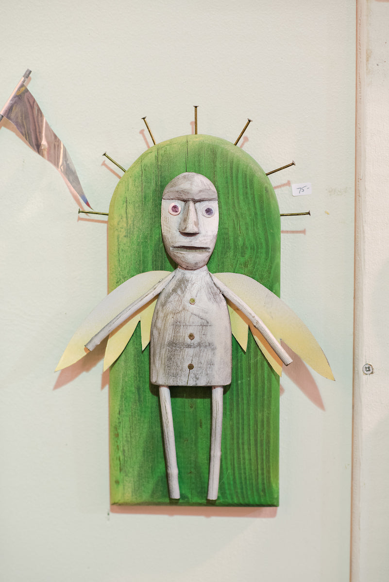 A wooden figure with wings and buttoned shirt, mounted on a green background with nails radiating like a halo, is displayed on a wall with a small price tag beside it.