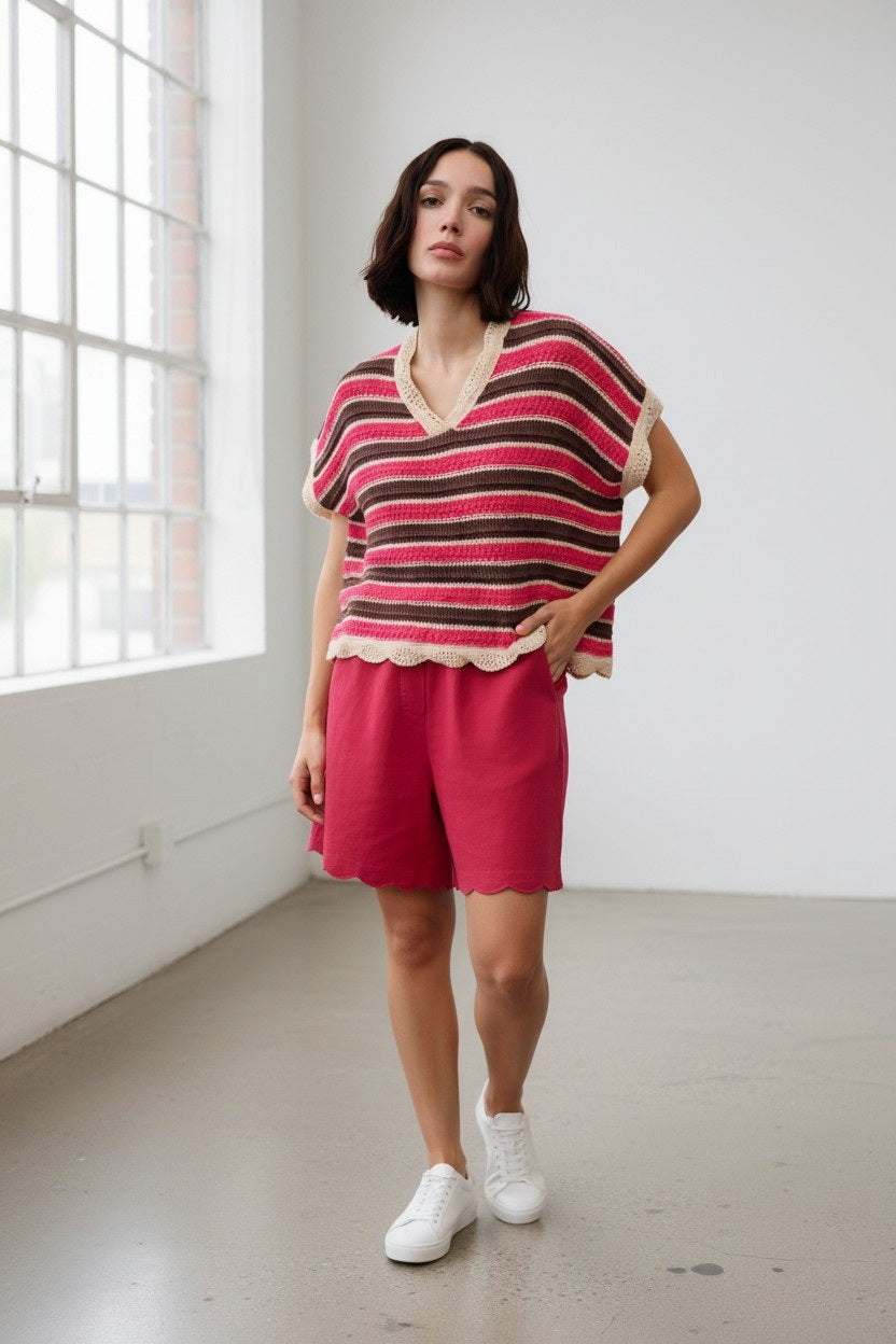 A woman stands indoors wearing the FLOOR - COTTON SWEATER WITH LACE DETAIL by FLOOR, paired with red shorts and white sneakers. She poses casually in a bright room with large windows and a concrete floor, embodying the effortless style of summer Italian knitwear.