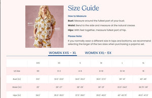 A woman with long, wavy brown hair stands barefoot against a white wall, wearing the PRINTFRESH Dachshund Date Short Sleeve Top & Cropped Pants Pajama Set in Blue. The organic cotton set from PRINTFRESH features short sleeves and cropped pants adorned with a charming dachshund print. She smiles slightly.