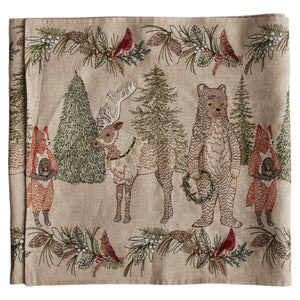A CHRISTMAS CHEER EMBROIDERED LINEN TABLE RUNNER featuring woodland animals in winter attire and Christmas trees, draped over a wooden dining table set with plates, glassware, and candlesticks.