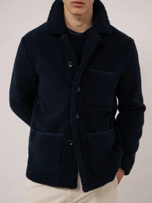 A young person with short dark hair wears the HARTFORD - Sherpa Utility Jacket in Navy by HARTFORD over a black shirt, looking directly at the camera against a plain light background. This warm sherpa layer brings both comfort and style to casual menswear.