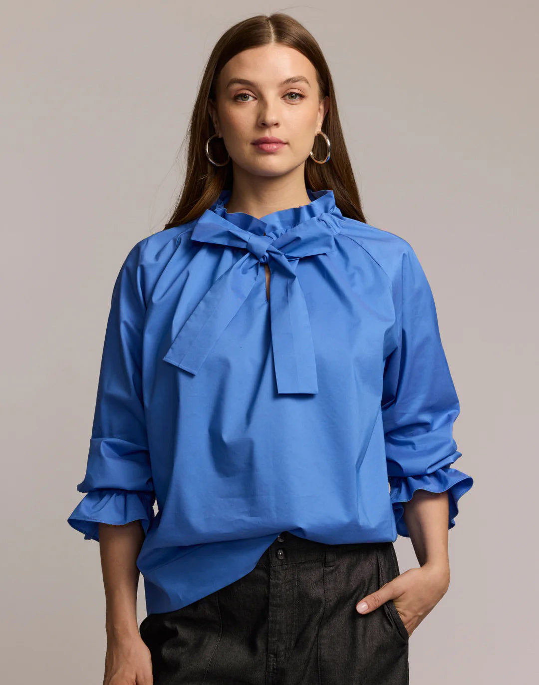 A woman with long brown hair models the HINSON WU ALINA LONG SLEEVE SHIRT, a bright blue blouse with a bow neck and ruffled sleeves, paired with dark pants. She stands against a light background, looking at the camera.