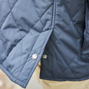 A man with light brown hair smiles while standing outside in a park. He wears the COLLARS AND CO - MIDDLEBURG QUILTED FIELD JACKET by COLLARS AND CO, featuring a corduroy collar, layered over a light blue shirt and tan pants for lightweight warmth on a cool day. Trees and a stone wall are visible in the background.