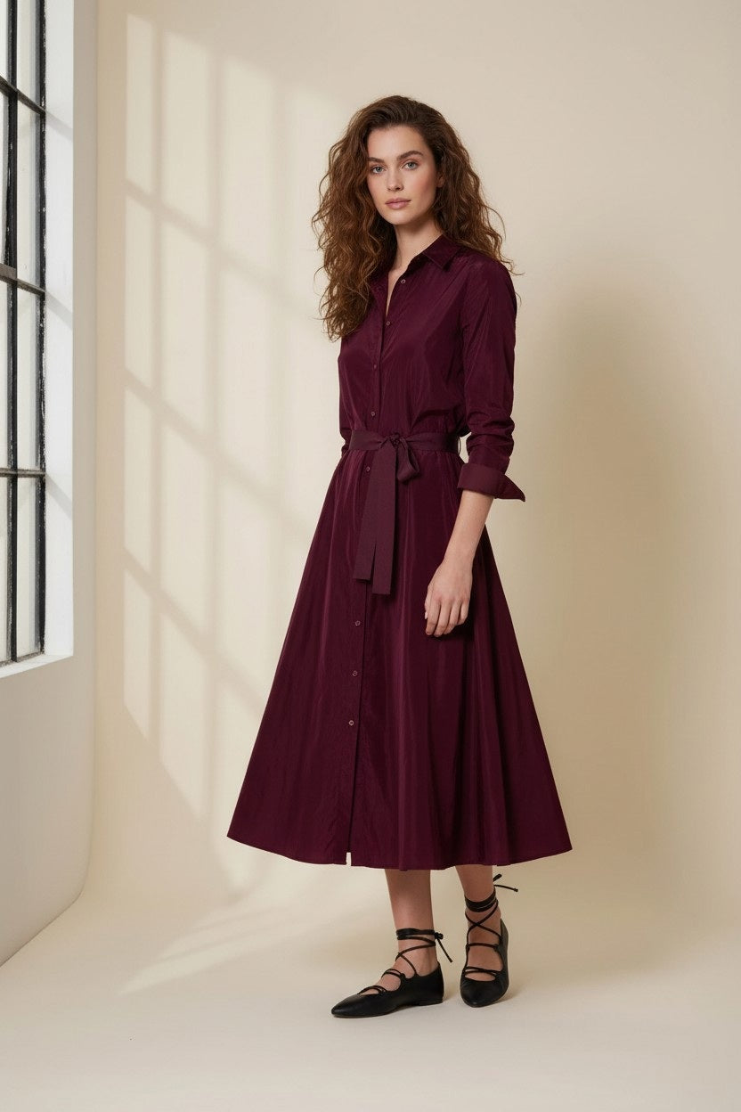 A person with long curly hair stands in a softly lit room by a window, wearing the VICARIO CINQUE ANNA Taffeta Shirtdress in Bordeaux and black lace-up flats.