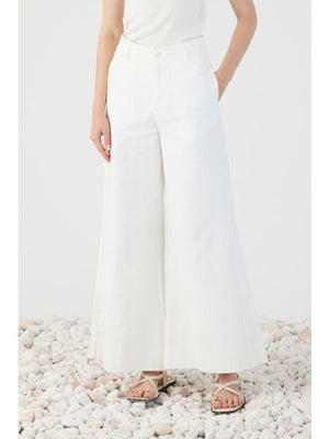 A person stands on white pebbles, shown from the back, wearing ELEH’s WIDE LEG COTTON TROUSER paired with a white sleeveless top and sandals—an effortless take on modern minimalism.