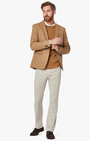 A bearded man stands against a plain background, wearing a tan blazer, a brown sweater over a white shirt, and 34 HERITAGE - CHARISMA RELAXED STRAIGHT LEG PANT IN DAWN TWILL. He adjusts his blazer sleeve with one hand.