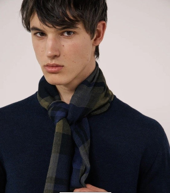 A young person with short dark hair wears a navy sweater and the HARTFORD Tartan Wool Scarf in Navy, Indigo, and Olive from HARTFORD, tied loosely around their neck, looking directly at the camera against a plain light background.