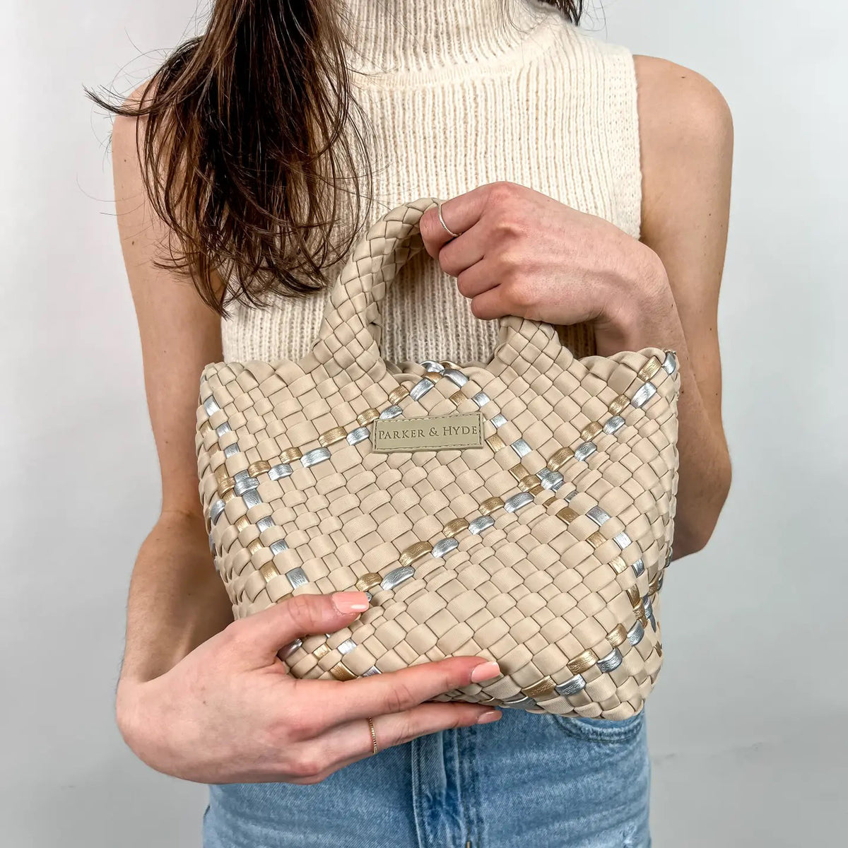 A woman with long brown hair, in a sleeveless cream knit top and blue jeans, holds a PARKER AND HYDE - MINI WOVEN TOTE BAG. The hand-woven tote by PARKER &amp; HYDE features a subtle checkered pattern with light blue accents.