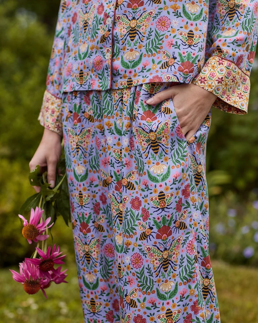 A person wearing the PRINTFRESH - BEE TAPESTRY ECO SATIN SOHO SET IN LAVENDER, featuring vintage-inspired bee and floral vine prints, holding a flower behind their back in a garden.