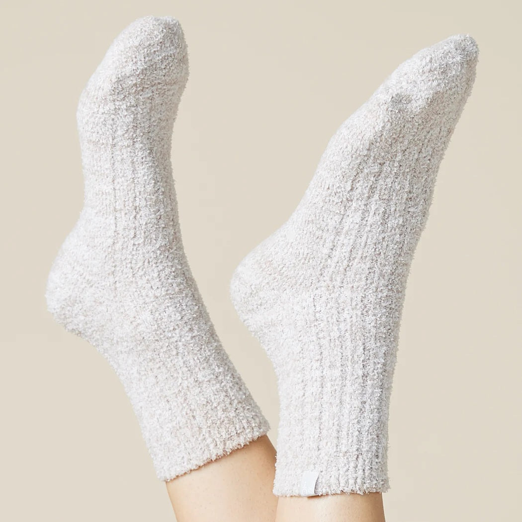 A pair of feet wear SOFTIES' Marshmalow Crew Socks in Heather Coco, raised against a beige background. These plush, cozy socks cover the ankles and add comfort with their soft, textured design.
