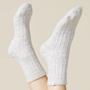 A pair of feet wear SOFTIES' Marshmalow Crew Socks in Heather Coco, raised against a beige background. These plush, cozy socks cover the ankles and add comfort with their soft, textured design.