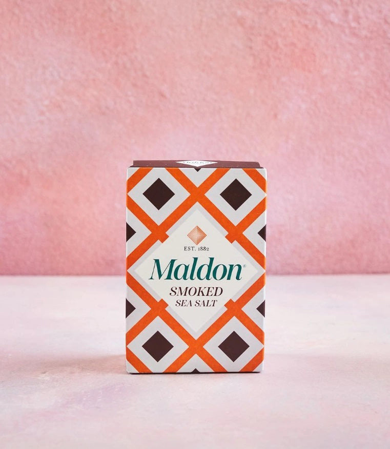 A box of GOURMET FOOD SOLUTIONS MALDON - SMOKED SEA SALT with an orange, brown, and white geometric design sits on a light pink surface against a textured pink background.