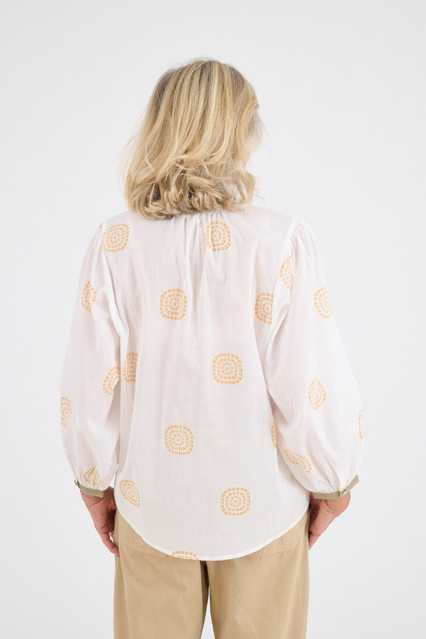 A smiling older woman with blonde hair stands against a white background, hands in her pockets, wearing the STORIATIPIC - LUCIE SOLEIL BLOUSE by STORIATIPIC, featuring orange patterns for a look of casual elegance.