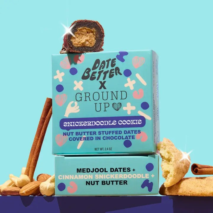 A box of DATE BETTER SNACKS’ DATE BETTER - SNICKERDOODLE COOKIE features nut butter stuffed dates enrobed in dark chocolate, artfully arranged with nuts, a cinnamon stick, and a bitten cookie to showcase the ultimate Snickerdoodle Cookie Crunch—all presented on a pastel backdrop.