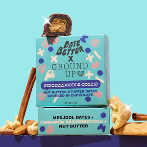 A box of DATE BETTER SNACKS’ DATE BETTER - SNICKERDOODLE COOKIE features nut butter stuffed dates enrobed in dark chocolate, artfully arranged with nuts, a cinnamon stick, and a bitten cookie to showcase the ultimate Snickerdoodle Cookie Crunch—all presented on a pastel backdrop.