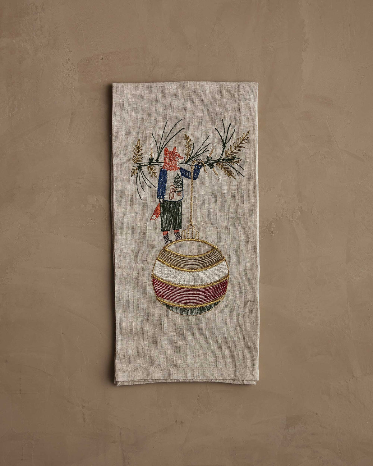 A beige linen DECK THE HALLS FOX LINEN TEA TOWEL embroidered with a fox holding a large, striped Christmas ornament, perfect for vintage Christmas kitchen decor.