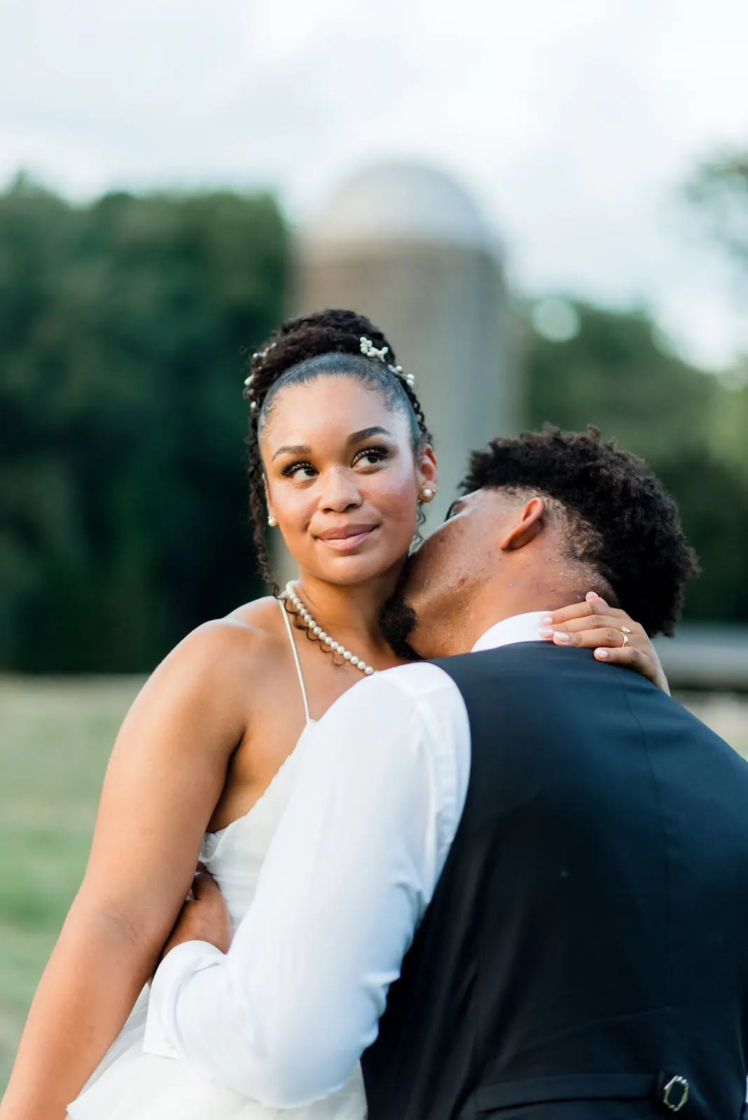 A bride in a white dress smiles softly while holding a groom in a dark vest, who embraces her and kisses her neck outdoors with blurred trees in the background.