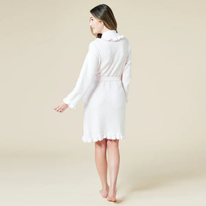 A woman with long hair stands barefoot against a light beige background, smiling softly in the SOFTIES - RUFFLE CHENILLE ROBE 36" IN PINK, featuring a belted waist and ruffled edges, exuding comfort and elegance.