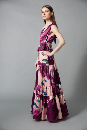A woman with straight brown hair and a neutral expression wears the HOPE FOR FLOWERS Tiered Maxi Dress, a sleeveless, floor-length style featuring bold purple and pink floral print, standing against a gray background.