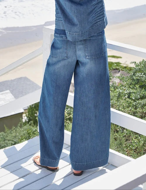 A person wearing FRANK & EILEEN’s Madeline Patch Pocket Pant in Famous Denim Distressed Wash stands on a white wooden deck overlooking greenery, sand, and the ocean, with only their lower body visible.