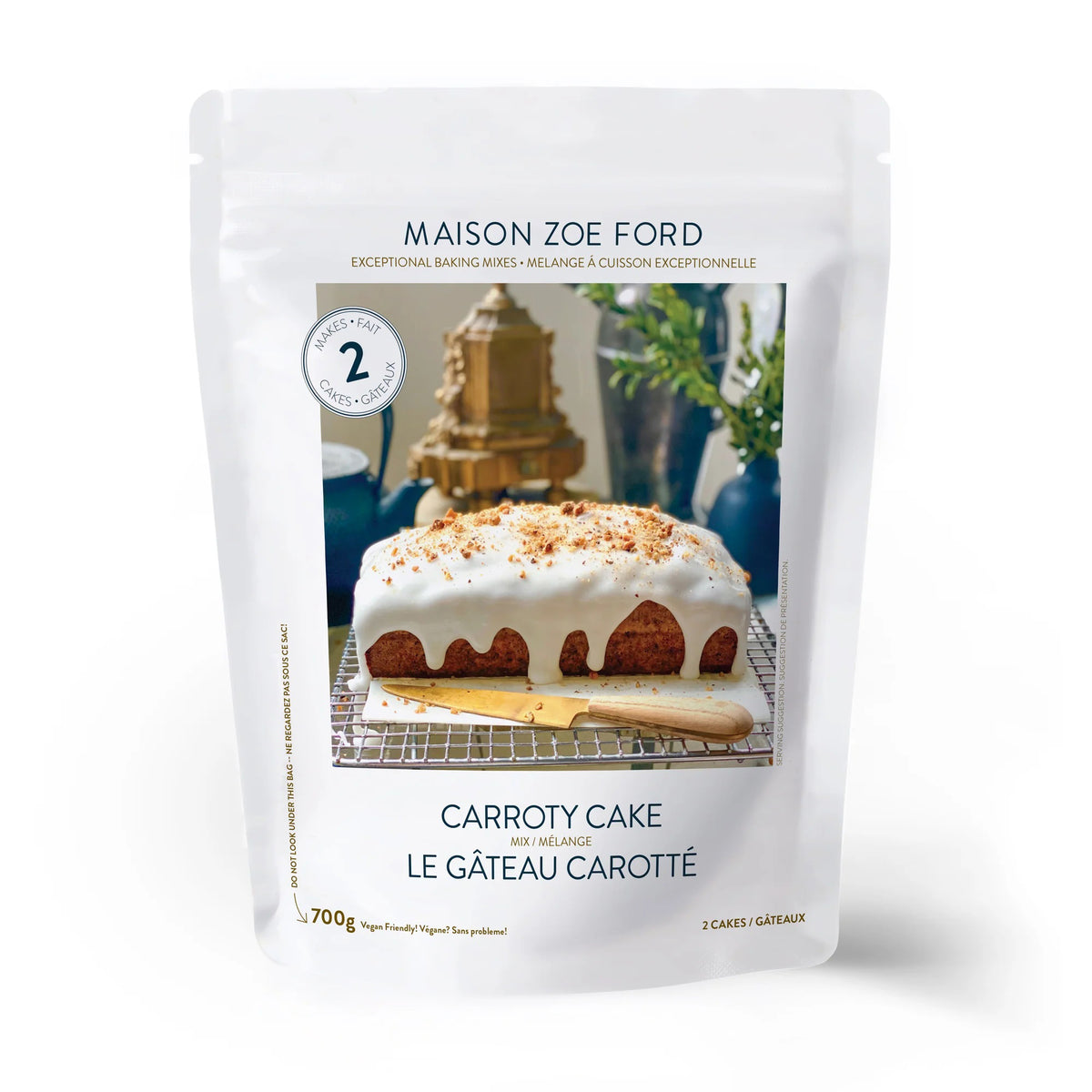 A package of MAISON ZOE FORD - CARROTY CAKE MIX is displayed, featuring an enticing image of a luscious carrot cake adorned with creamy white icing and sprinkled with nuts, making it an ideal addition to any dessert table. The text reads "Exceptional Baking Mixes" and "700g." The predominantly white packaging exudes a sense of simplicity and elegance.