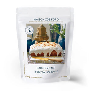 A package of MAISON ZOE FORD - CARROTY CAKE MIX is displayed, featuring an enticing image of a luscious carrot cake adorned with creamy white icing and sprinkled with nuts, making it an ideal addition to any dessert table. The text reads "Exceptional Baking Mixes" and "700g." The predominantly white packaging exudes a sense of simplicity and elegance.